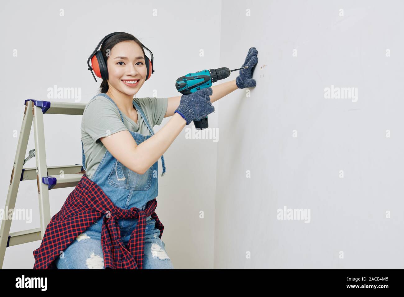 Portrait of pretty smiling young Asian woman wearing protective ear muffs when using electric drill Stock Photo