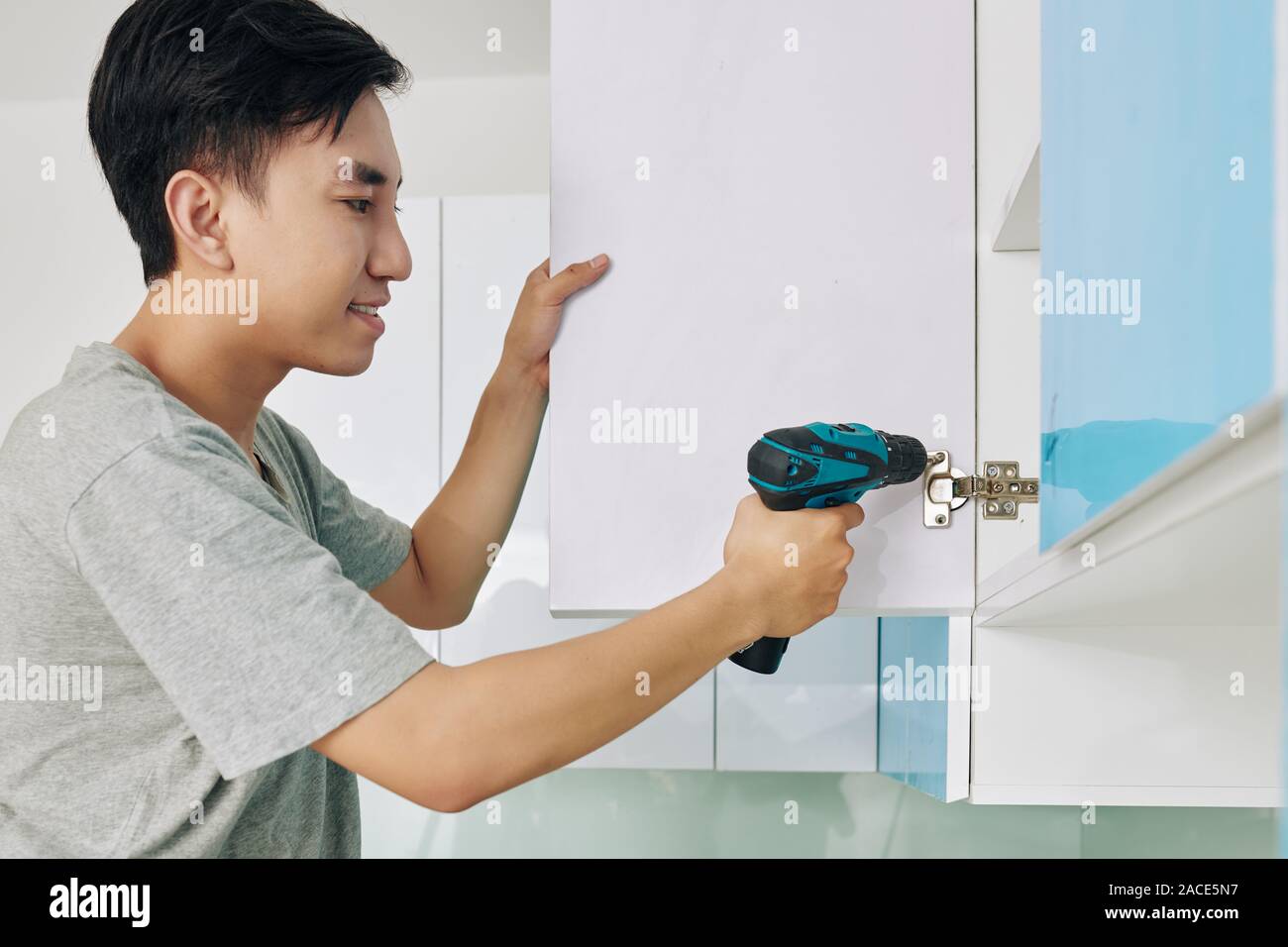 Handsome young Vietnamese man using electric screwdriver when assembling cupboard in kitchen Stock Photo