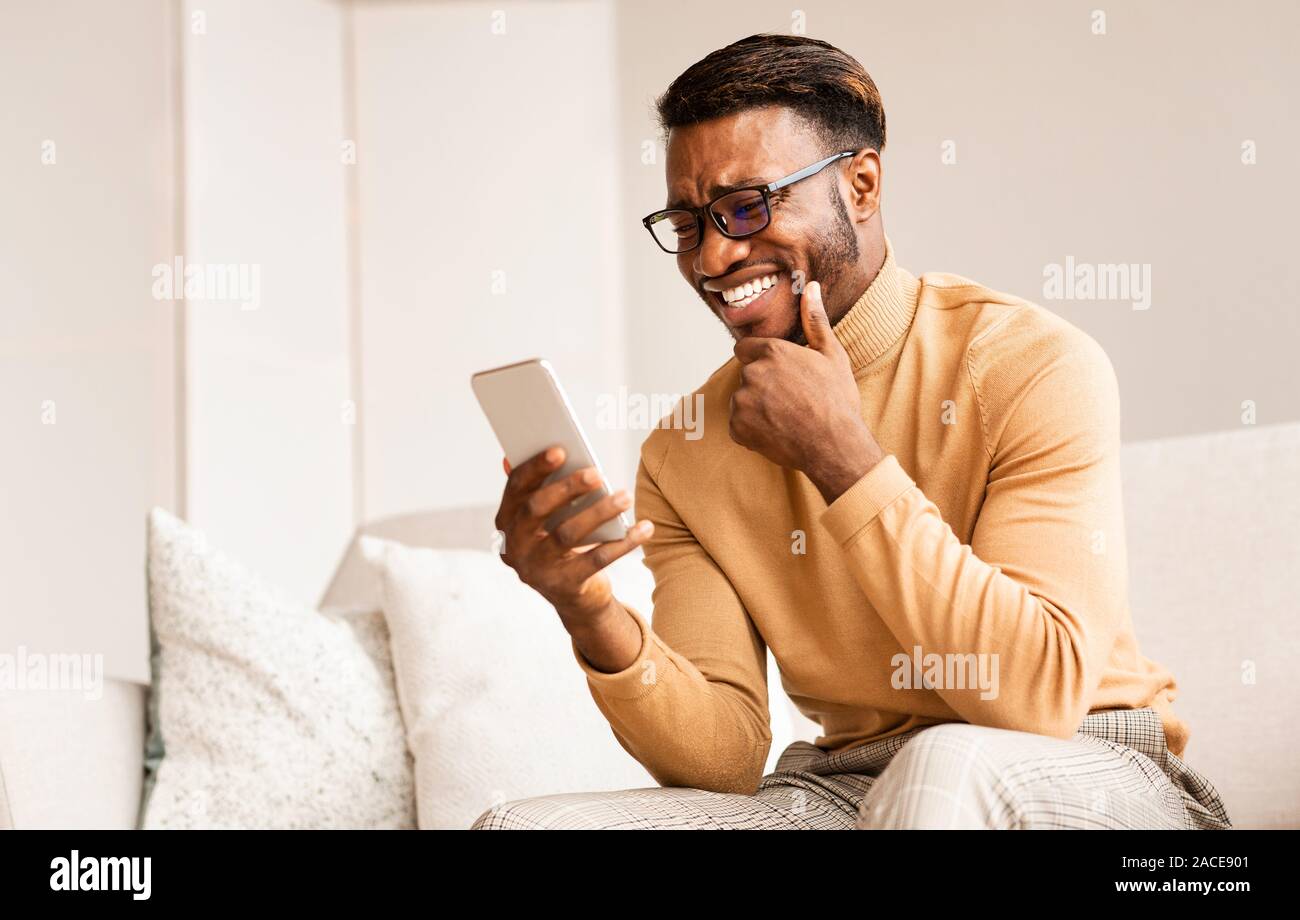 Afro Guy Using Cellphone Reading Funny News Sitting At Home Stock Photo
