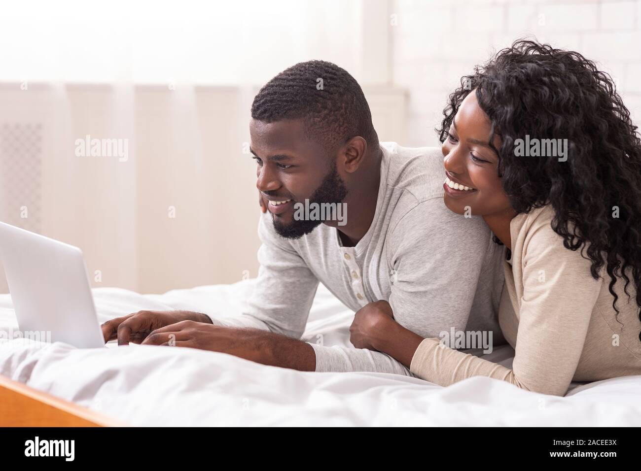 Cheerful afro spouses lying in bed using laptop together Stock Photo