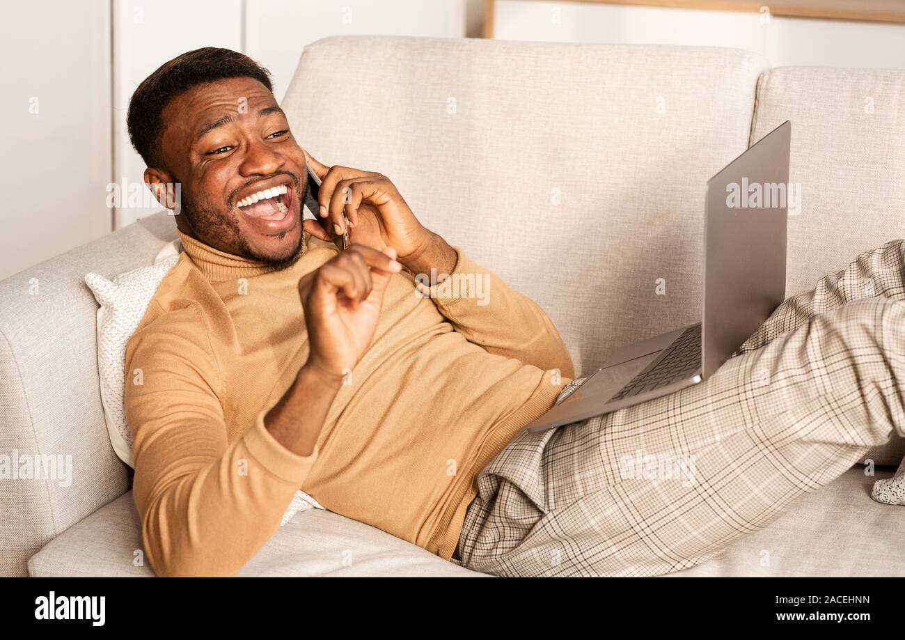 Cheerful Guy Talking On Phone Lying With Laptop On Couch Stock Photo