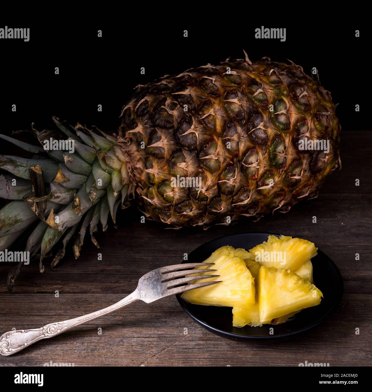 Sliced pineapple on old rustic desk with knife and with whole pineapple behind. Black background. Stock Photo
