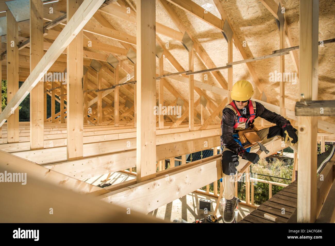 House Wooden Roof Skeleton Building and Caucasian Construction Contractor Worker Seating on a Beam. Building Technologies. Industrial Theme. Stock Photo