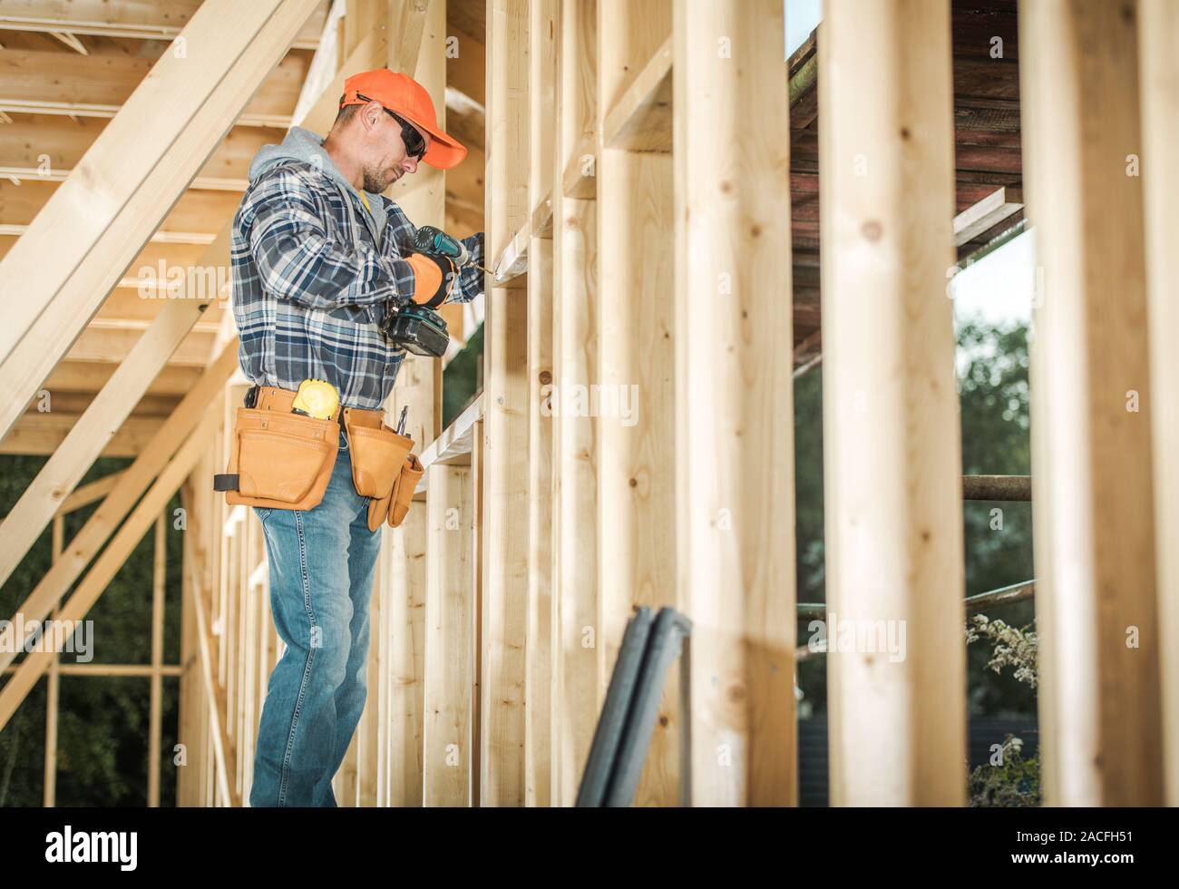 Wood House Frame Construction and the Contractor Worker in His 30s with Drill Driver Attaching Wooden Frame Elements. Industrial Theme. Stock Photo