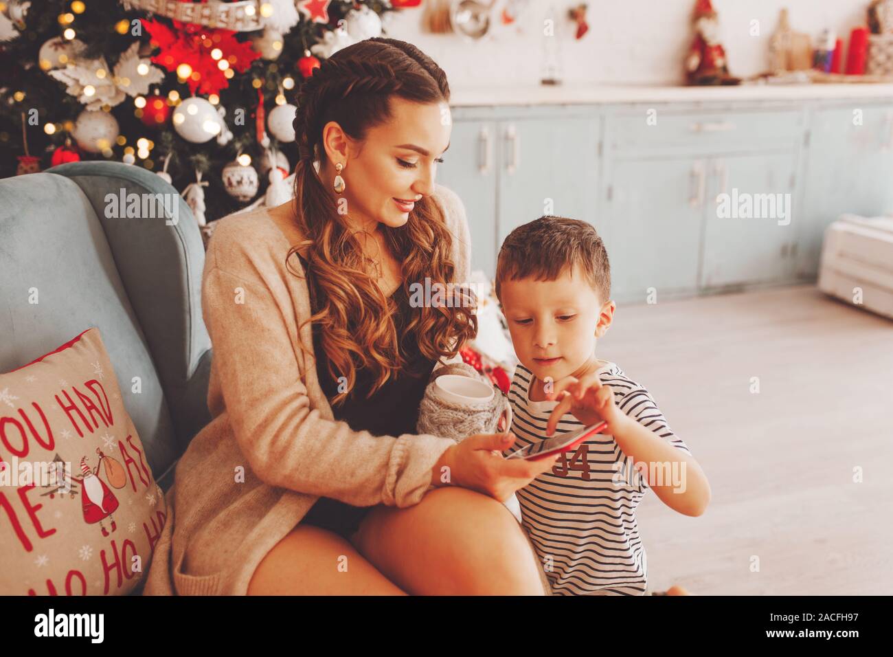 https://c8.alamy.com/comp/2ACFH97/young-mother-with-her-son-in-the-new-years-kitchen-near-the-christmas-tree-chooses-gifts-on-the-phone-she-holds-a-cup-of-coffee-or-tea-2ACFH97.jpg