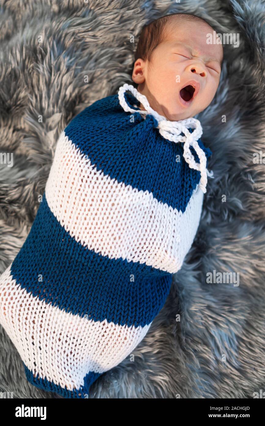 newborn baby boy yawning and swaddled in a knit wrap on a bed Stock