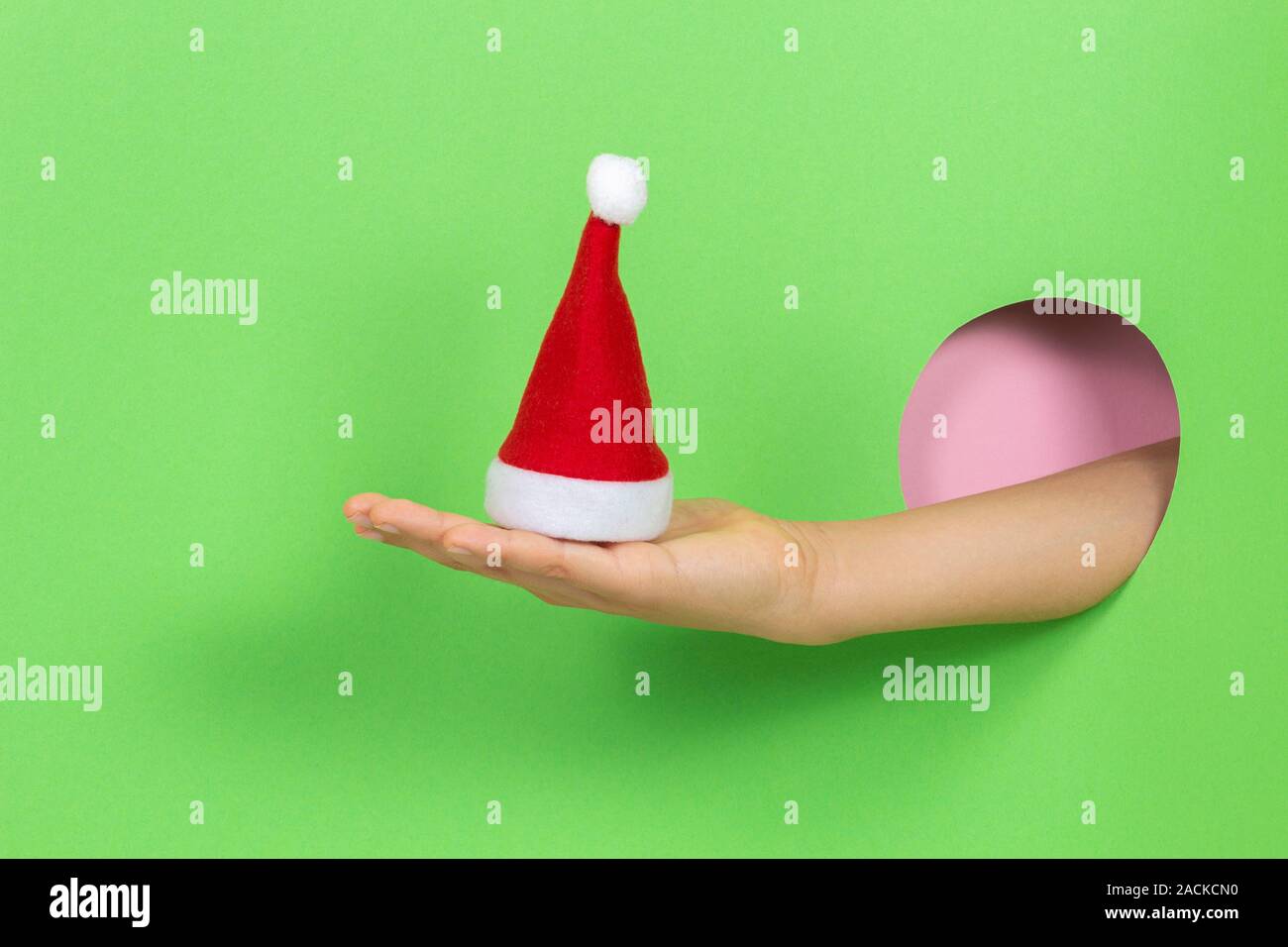 Hand through hole in light green background holding small red Christmas Santa Claus hat Stock Photo