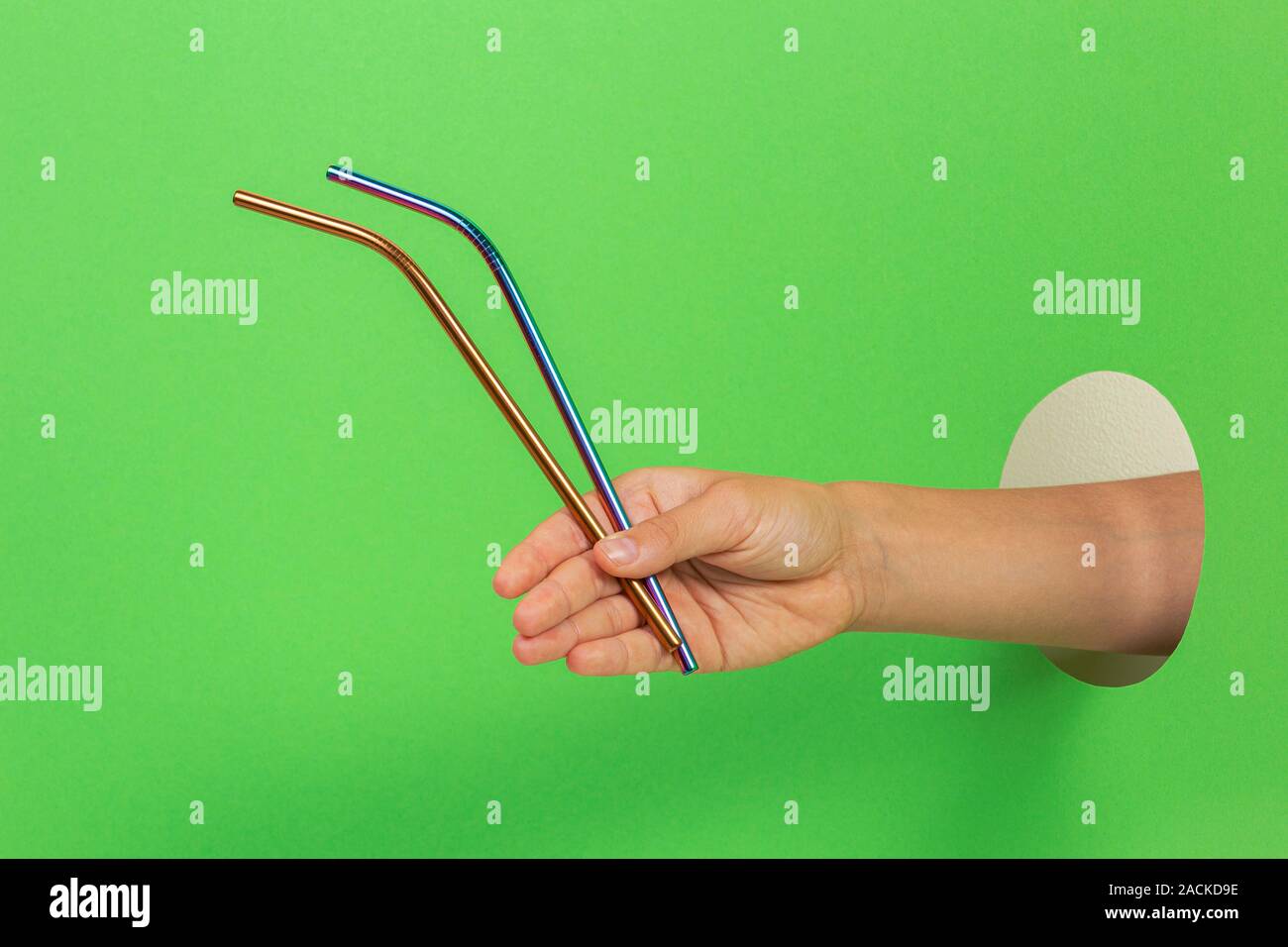 Hand through hole on light green background holding metallic reusable straws Stock Photo