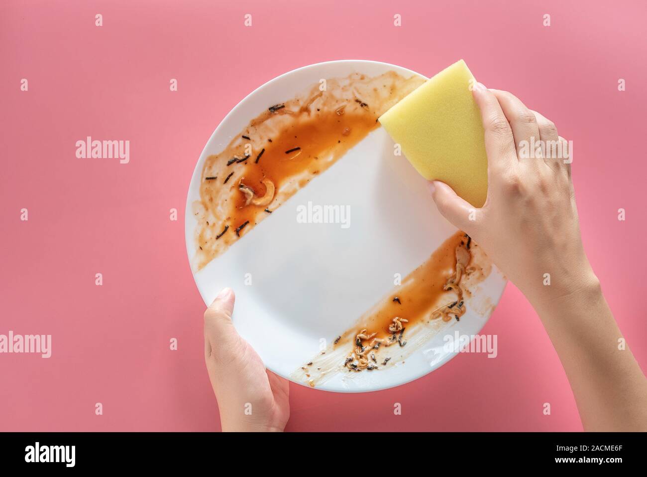 woman using yellow cleaning sponge to clean up and washing food stains and dirt on white dish after eating meal isolated on pink background. cleaning Stock Photo