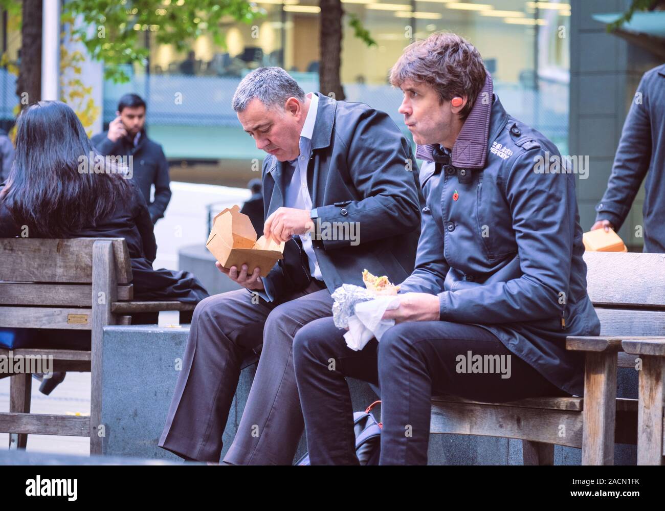 Two male city centre workers eating lunch outside on square bench wearing warm clothing. London City, UK Stock Photo
