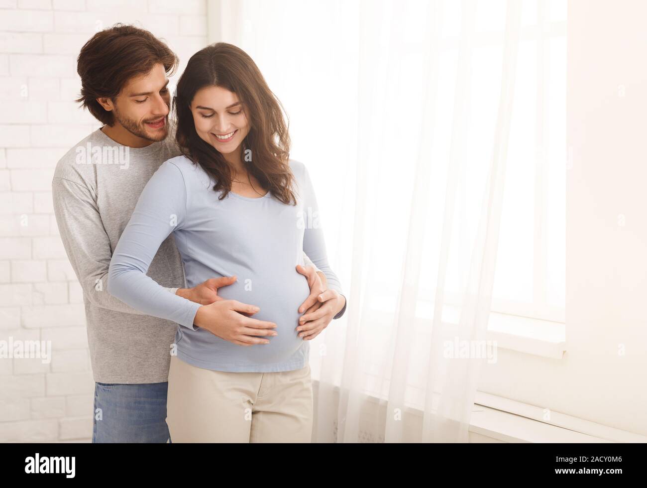 Loving man hugging his pregnant wife from behind standing near window ...