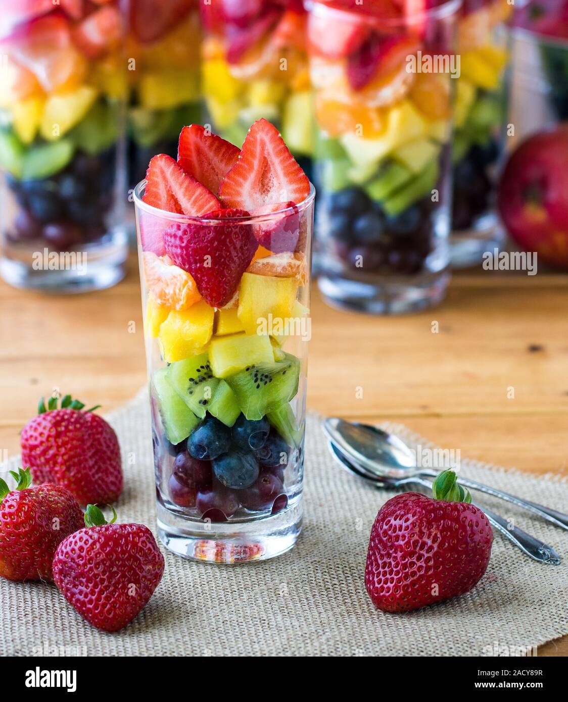 A close up view of a rainbow fruit parfait in a tall glass surrounded by strawberries and several rainbow parfaits in the background. Stock Photo