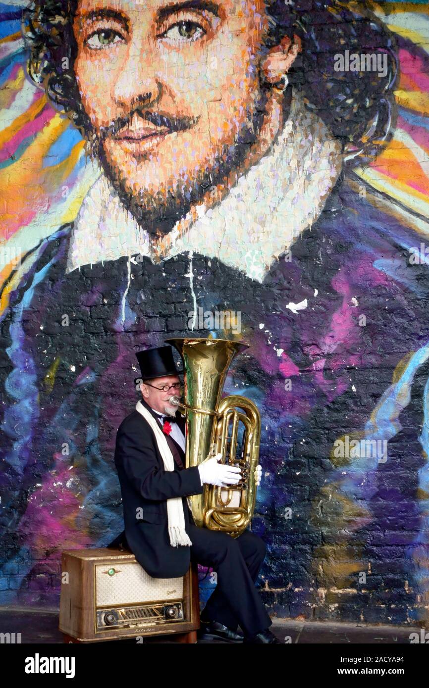 Tuba player street performer sitting on an amplifier at the base of a Shakespeare mural in a railway arch near the Globe Theatre, London Stock Photo