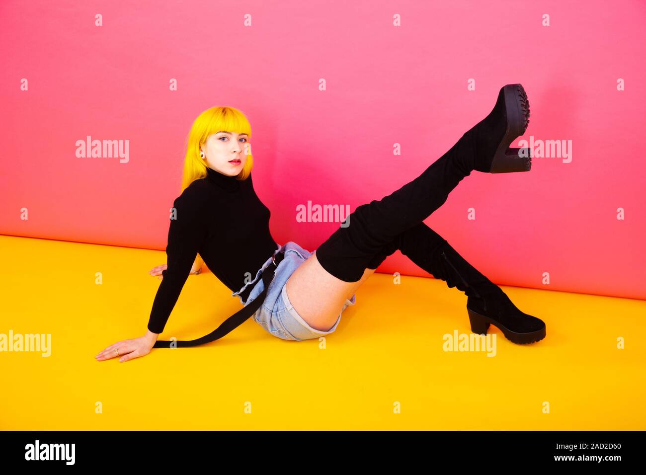 A young woman with bright yellow hair sitting down wearing over knee boots against a bright pink / yellow background Stock Photo