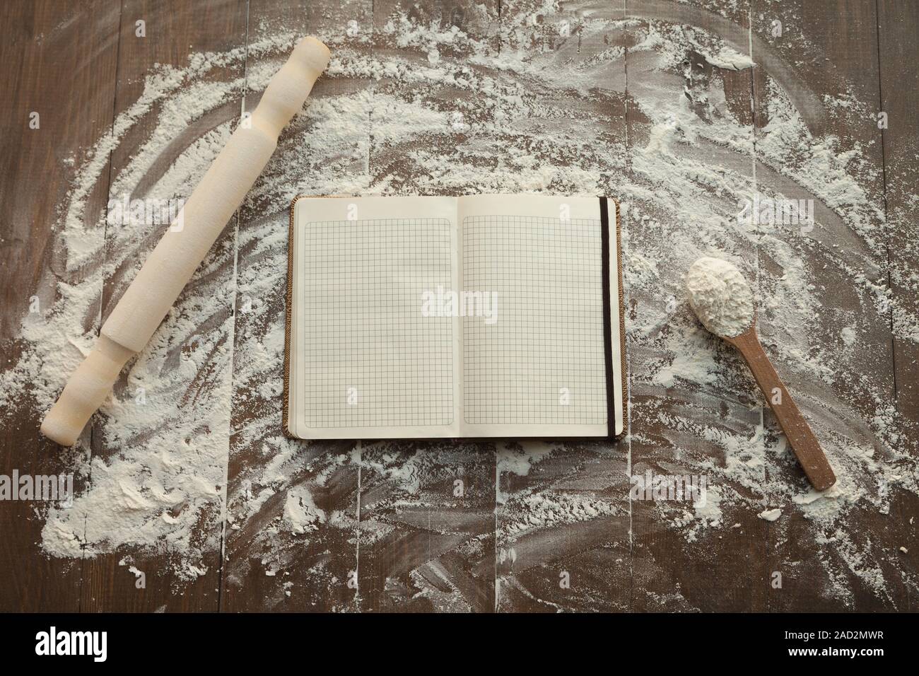 Composition of clear open recipe book in middle and rolling pin with spoon by sides on floured table. Stock Photo