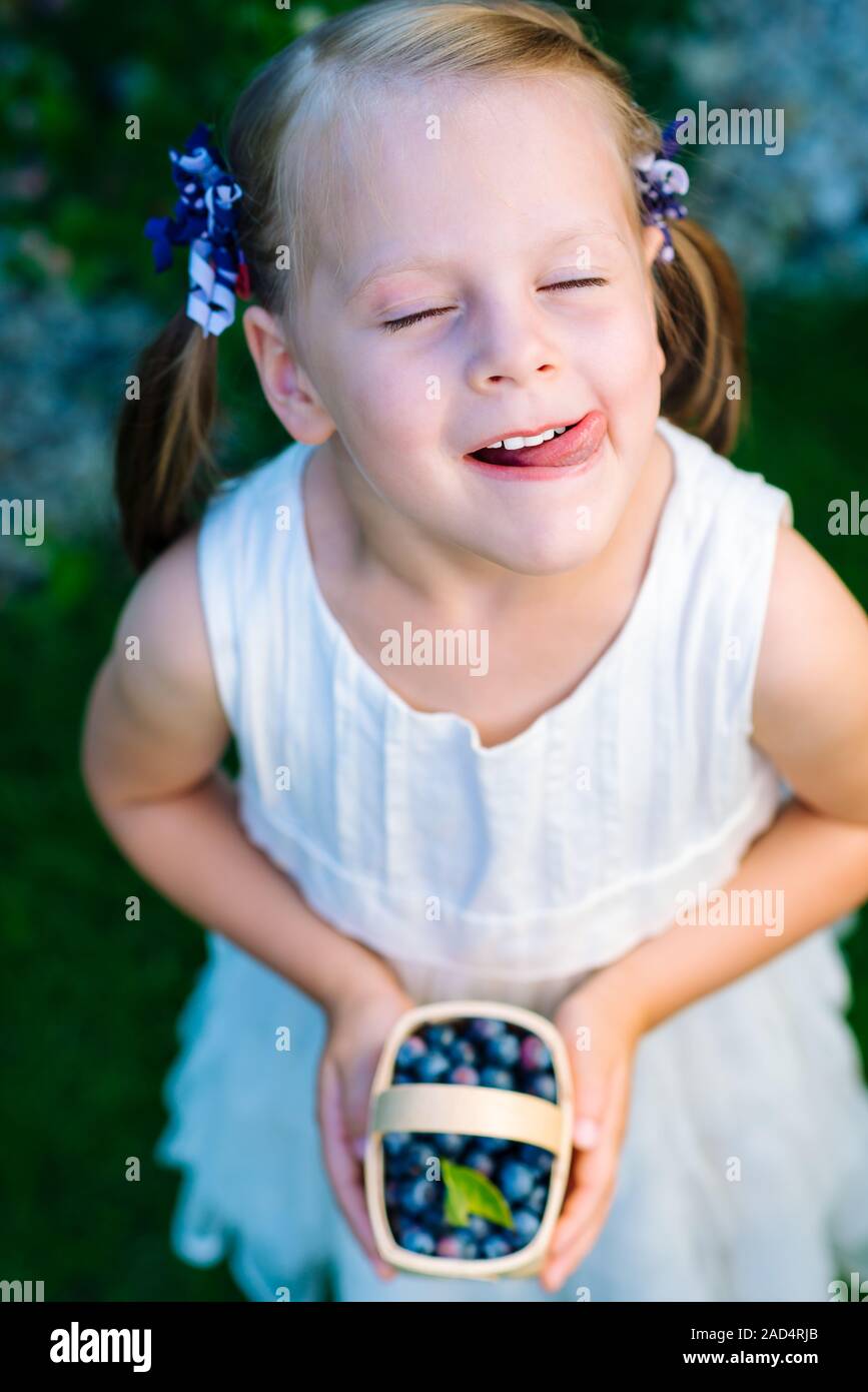 Little girl licking her lips with her eyes closed holding a basket of blueberries Stock Photo