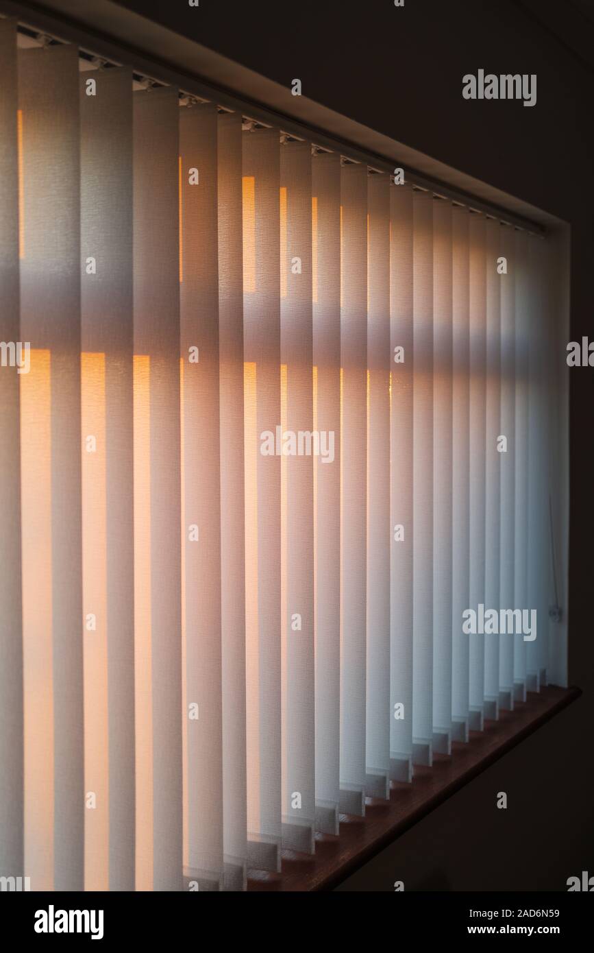 White vertical slat blinds hanging in front of a window as the sun is