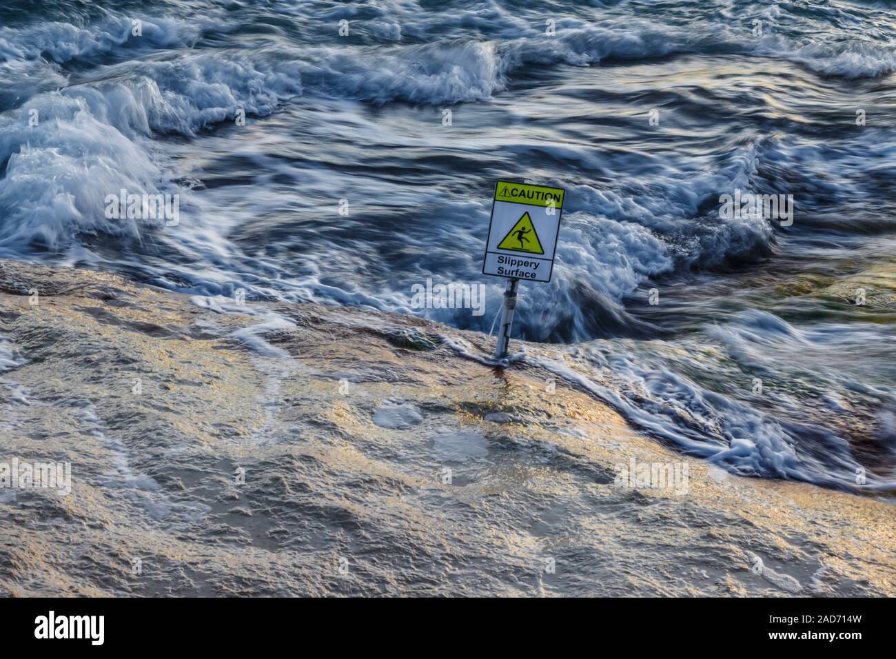 The sea swirls around a natural rock promenade with a sign warning of a slippery surface. Stock Photo