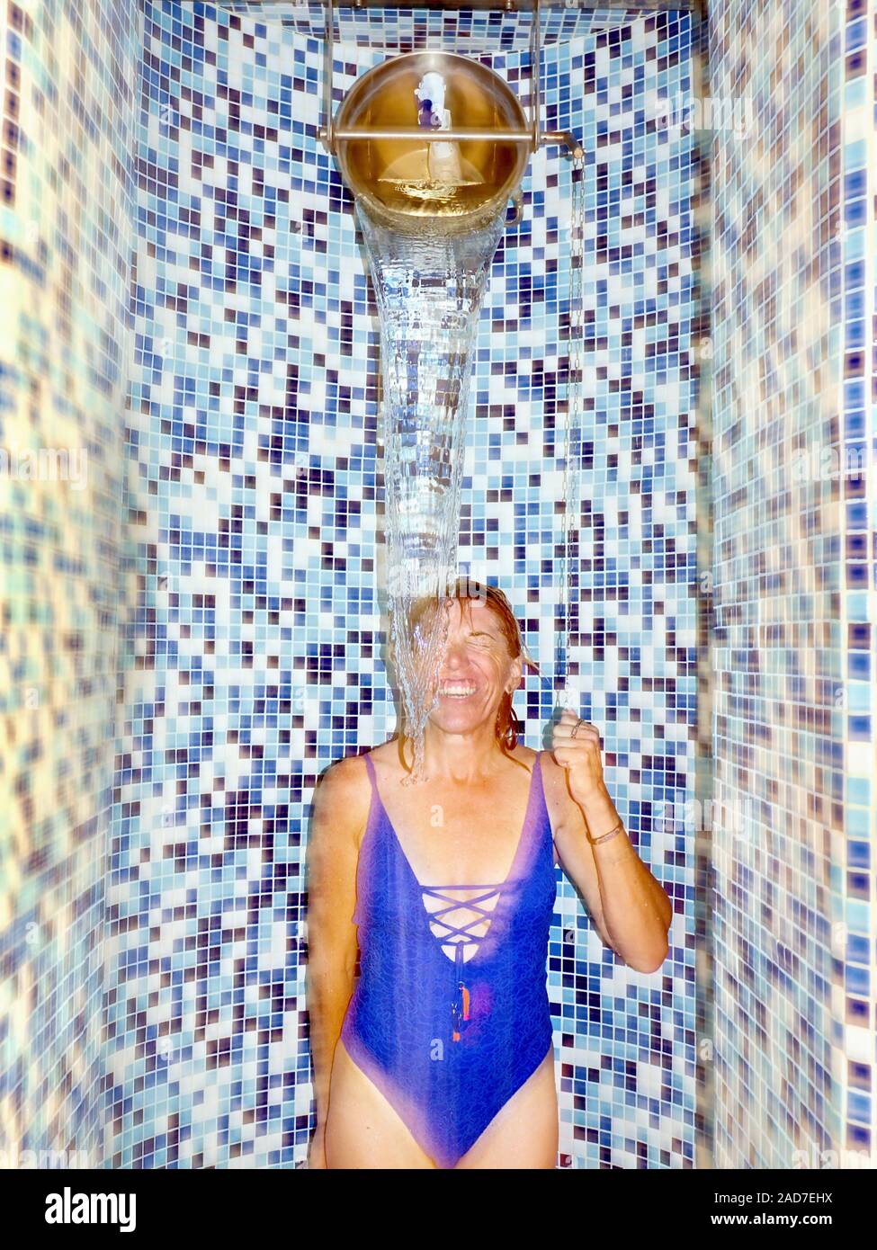 Wellness area with blue and white mosaics, a woman in a blue swimsuit standing under a pouring ice water bucket. Stock Photo