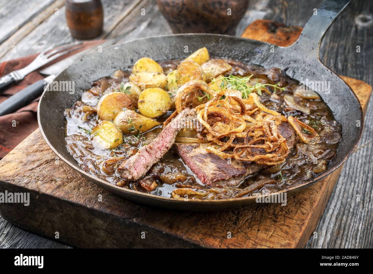 Traditional dry aged sliced roast beef with fried onion rings and potato chips as closeup in a wrough-iron pan with brown sauce Stock Photo