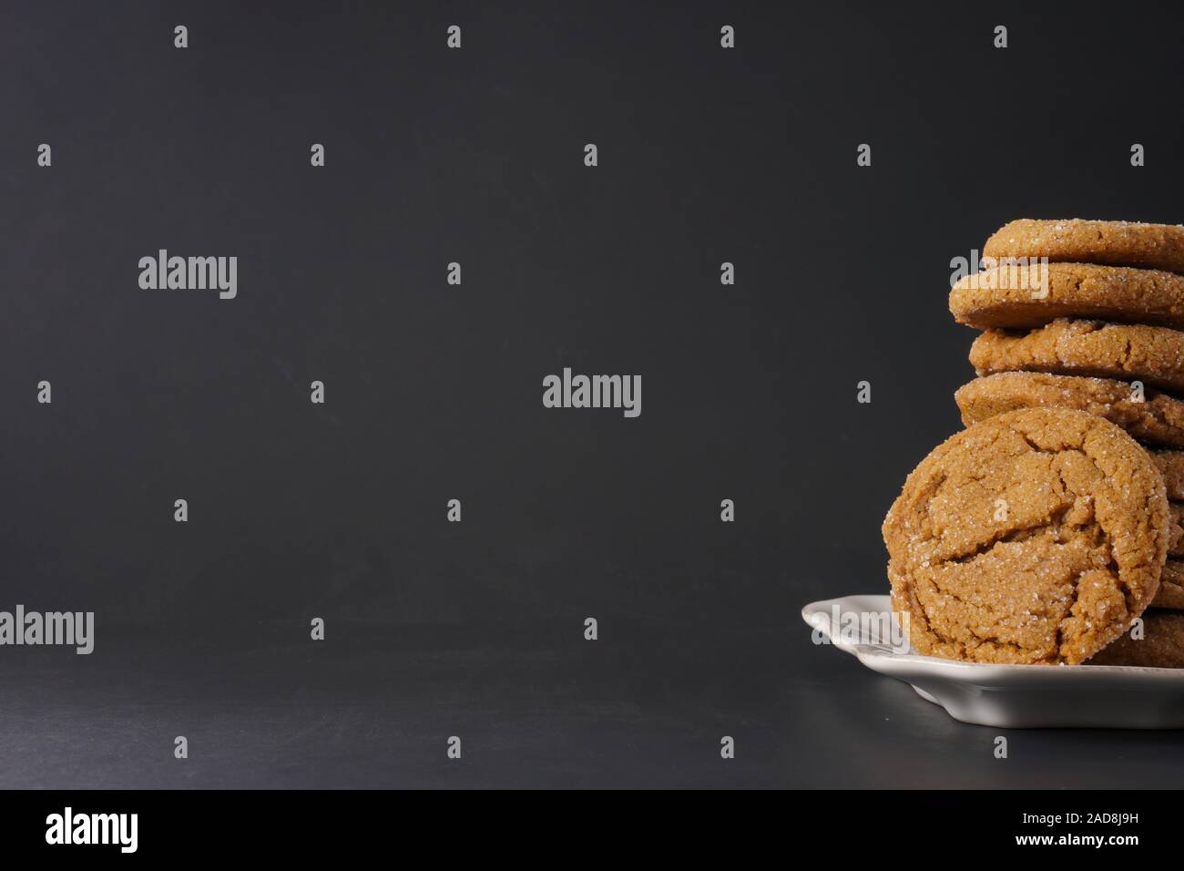 Partial view of a gingerbread cookie leaning against a stack of cookies on a white plate with a black background and copy space; landscape view Stock Photo