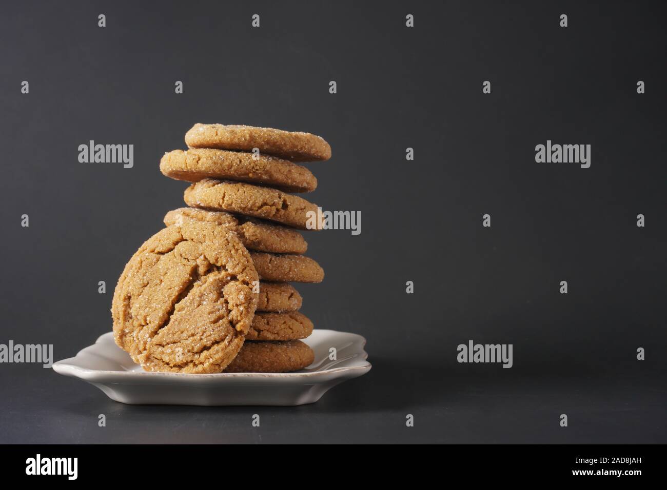 A crackly gingerbread cookie leans against a stack of cookies on a white plate with black background; copy space Stock Photo