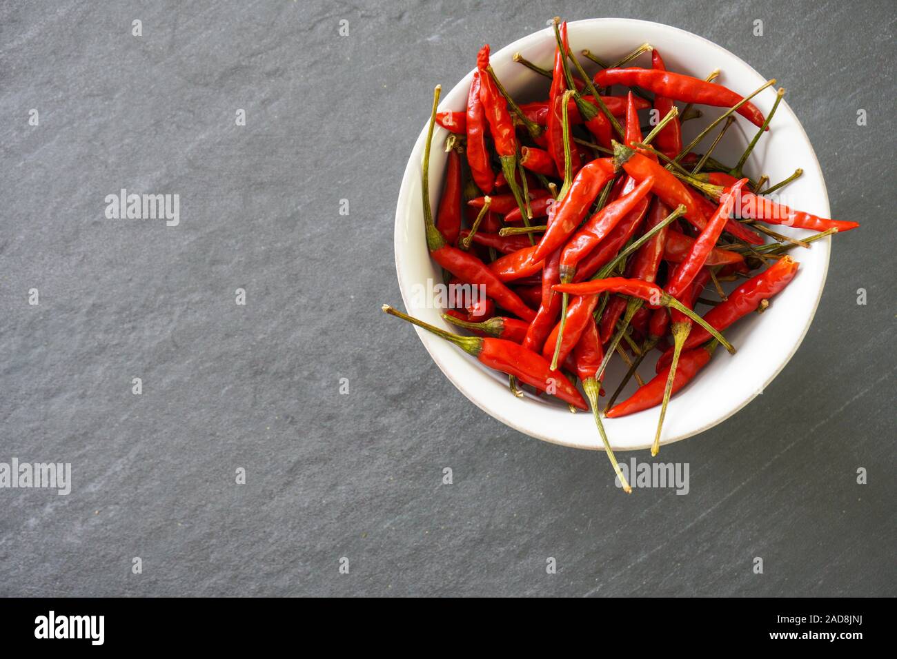 A white bowl full of bright red Thai chiles with green stems resting on a gray slate background with copy space; food and cooking Stock Photo