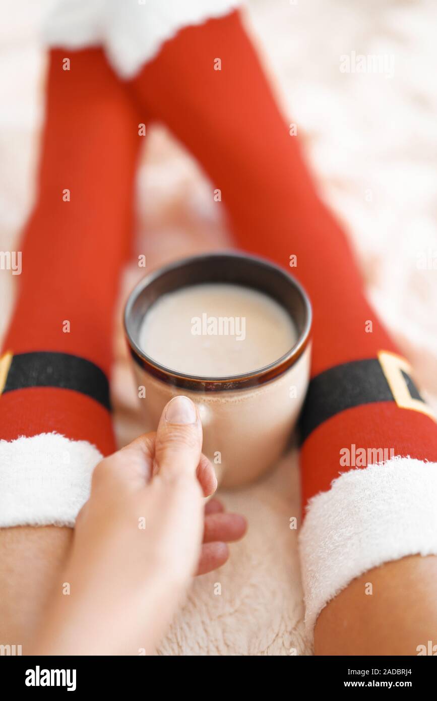 Feet in Santa's socks near the Christmas tree. Woman sitting at the blanket, drinks hot beverage and relaxes  warming up their feet in woollen socks. Stock Photo