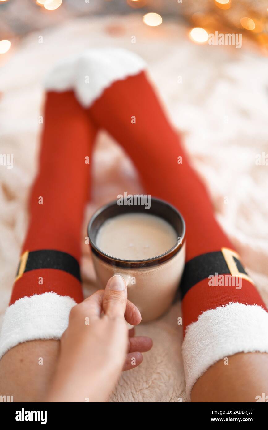 Lifestyle home photo of feet in Santa's socks near the Christmas tree. Woman sitting at the blanket, drinks hot beverage and relaxes  warming up their Stock Photo