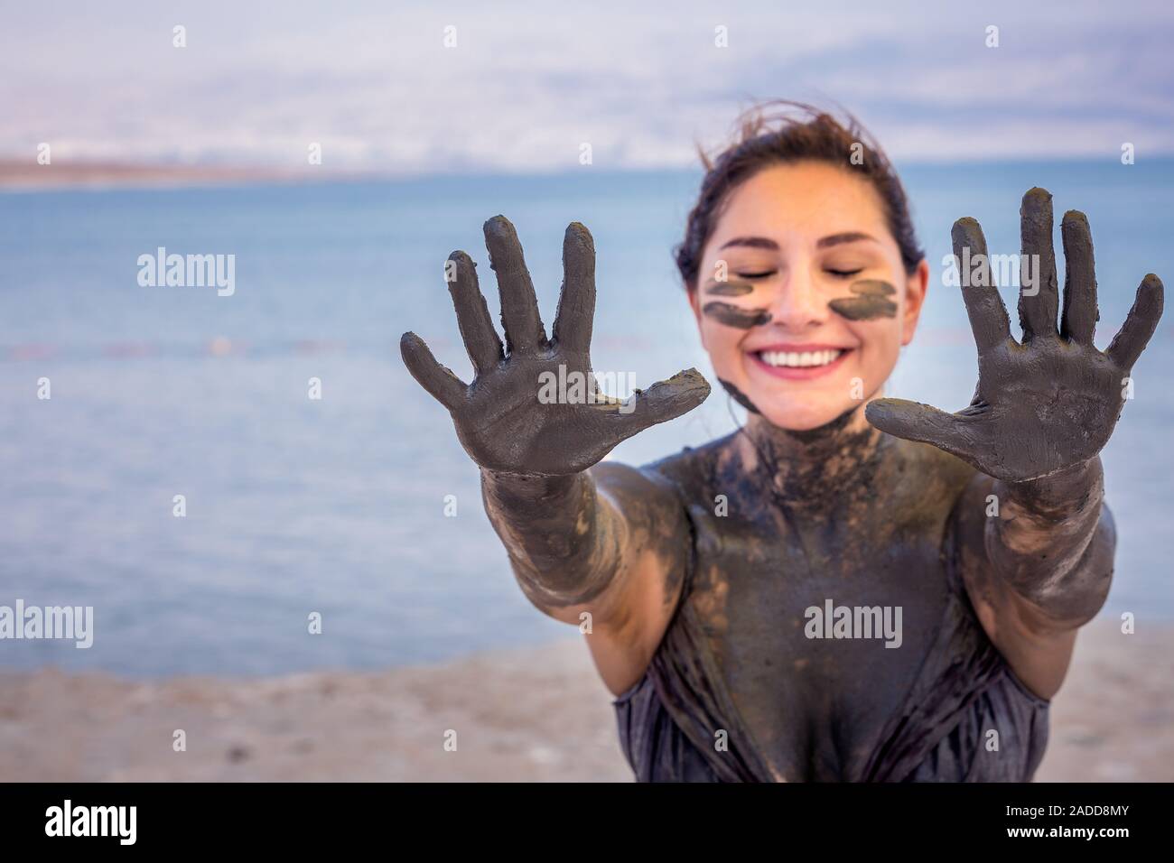 Happy smiling girl soiled in the mud of the Dead Sea shows her palms in the mud at Dead Sea, Israel. Copypaste space. Selective focus on hands. Stock Photo