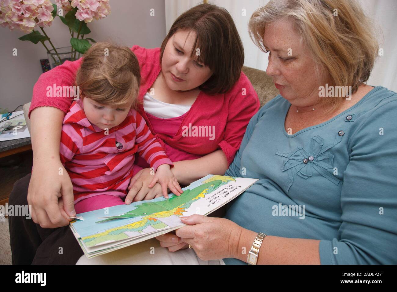 Toddler with her mum and grandmother on a sofa reading a book Stock