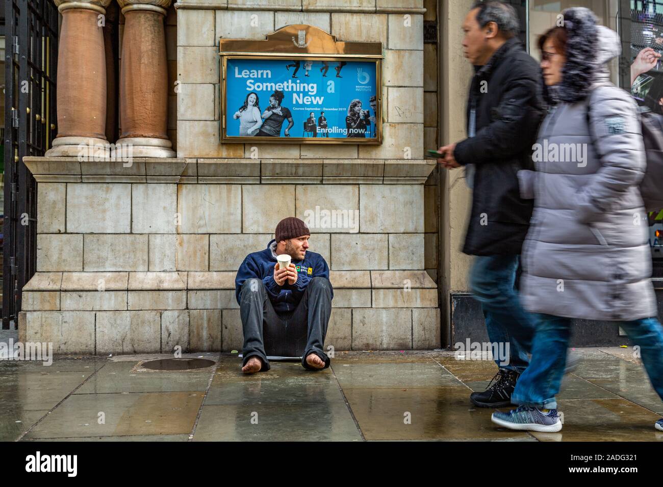 Man without shoes sitting down begging for money in winter, as people rush pass without caring, close to Liverpool Street station London England UK Stock Photo