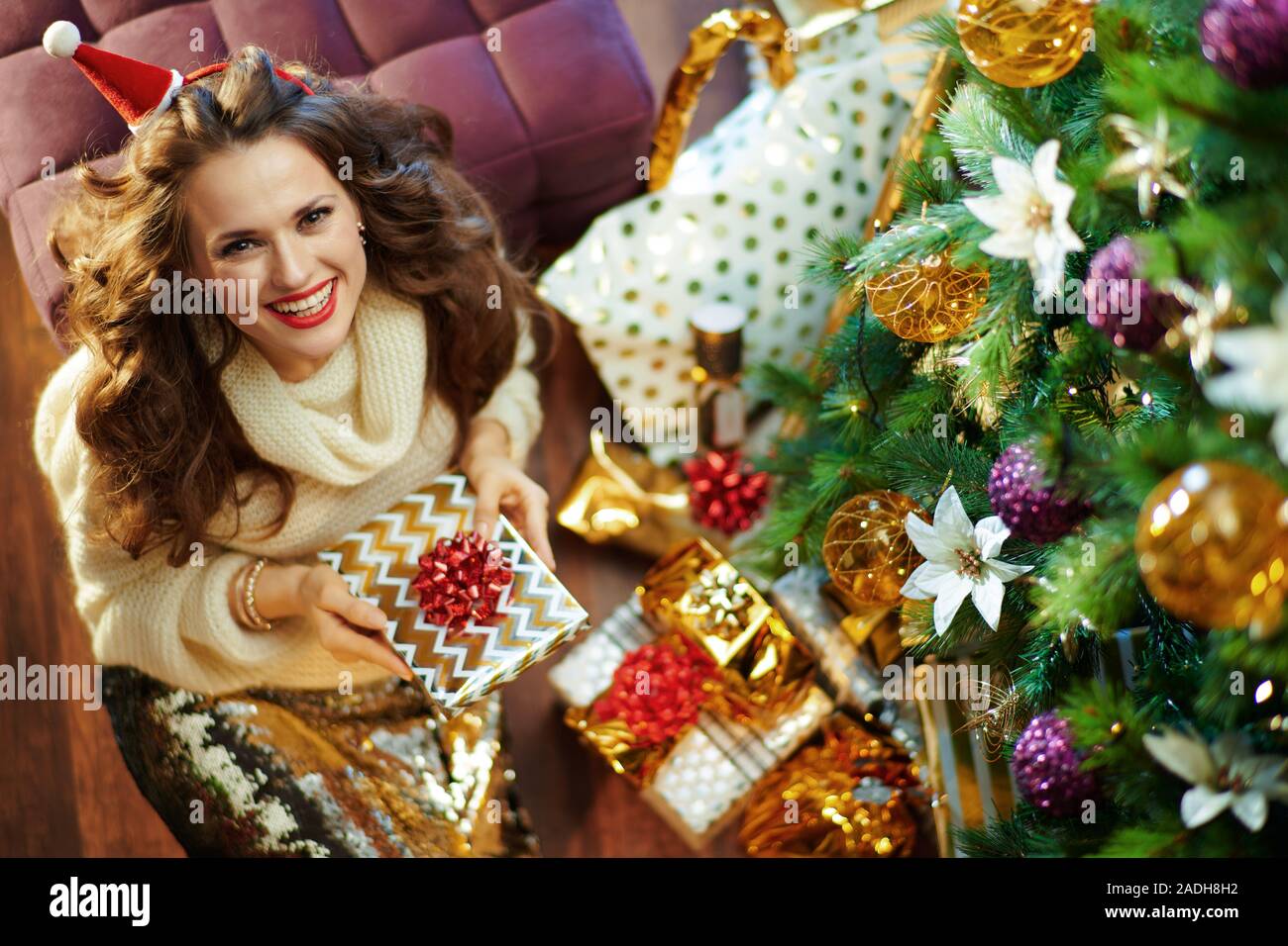 Upper view of smiling stylish woman with long brunette hair in gold sequin skirt and white sweater under decorated Christmas tree near present boxes h Stock Photo