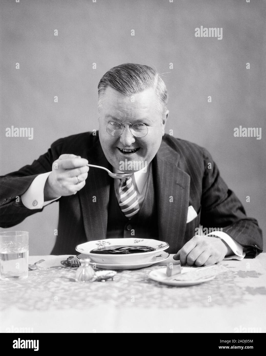 1930s MIDDLE-AGED MAN WEARING PINCE-NEZ EYEGLASSES PINSTRIPED SUIT LOOKING AT CAMERA SITTING AT DINING TABLE EATING BOWL OF SOUP - f5662 HAR001 HARS COPY SPACE HALF-LENGTH PERSONS MALES CONFIDENCE EYEGLASSES VEST EXPRESSIONS MIDDLE-AGED B&W MIDDLE-AGED MAN EYE CONTACT SUIT AND TIE HUNGRY HAPPINESS CHEERFUL ENJOYING NUTRITION ENTHUSIASTIC PINCE-NEZ SMILES SUPPER CONNECTION THREE PIECE SUIT CONSUME CONSUMING JOYFUL NOURISHMENT STYLISH PINSTRIPED EAGER BLACK AND WHITE CAUCASIAN ETHNICITY HAR001 OLD FASHIONED Stock Photo