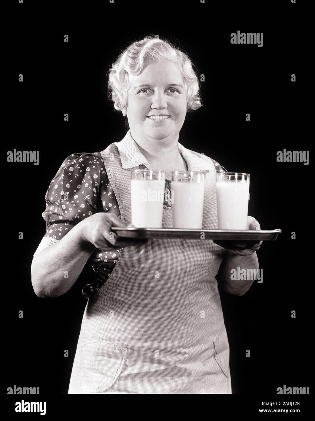 1940s SMILING MIDDLE-AGED WOMAN WEARING FARMHOUSE APRON LOOKING AT CAMERA CARRYING A TRAY WITH THREE GLASSES FULL OF MILK - f9538 HAR001 HARS PERSONS CARING CONFIDENCE MIDDLE-AGED B&W EYE CONTACT FARMHOUSE HAPPINESS MIDDLE-AGED WOMAN CHEERFUL PRIDE SMILES WHITE HAIR JOYFUL GRAY HAIR BLACK AND WHITE CAUCASIAN ETHNICITY HAR001 OLD FASHIONED Stock Photo