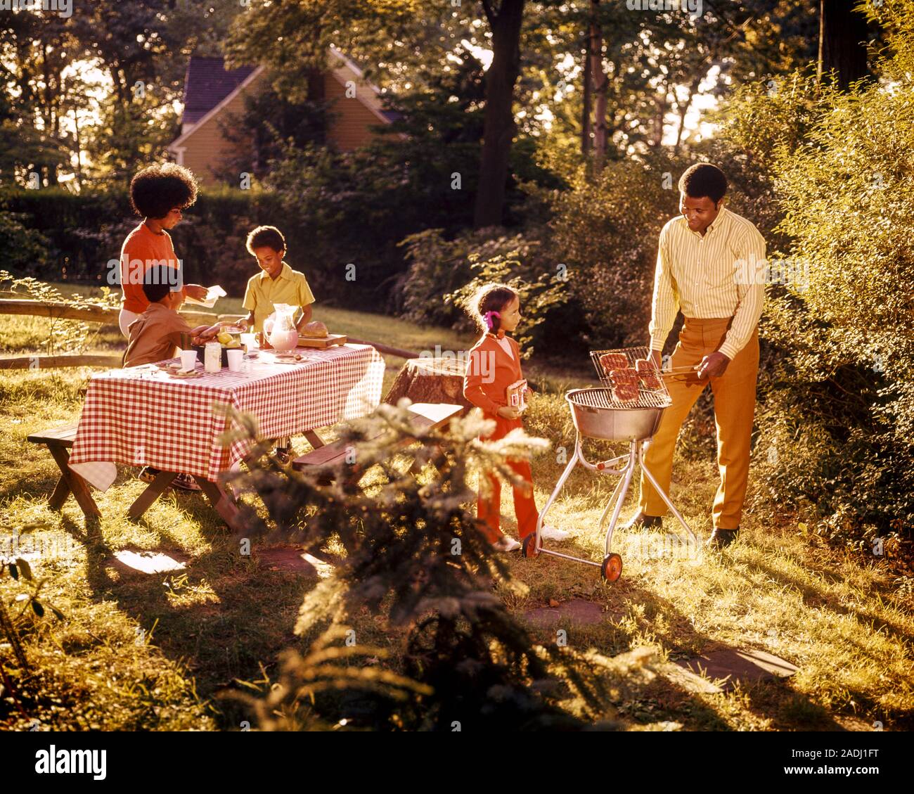1970s AFRICAN AMERICAN FAMILY HAVING BBQ PICNIC IN BACKYARD DAD