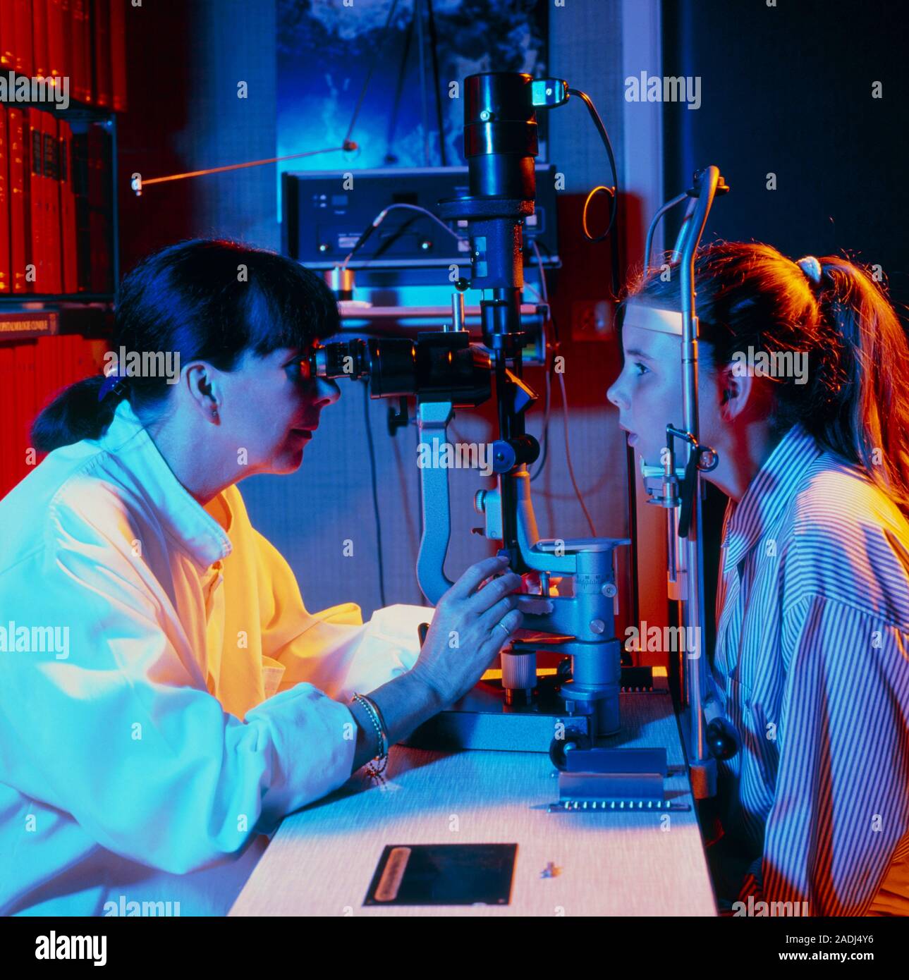 Eye examination. Optician uses an ophthalmoscope to examine the eyes of a young girl. At left