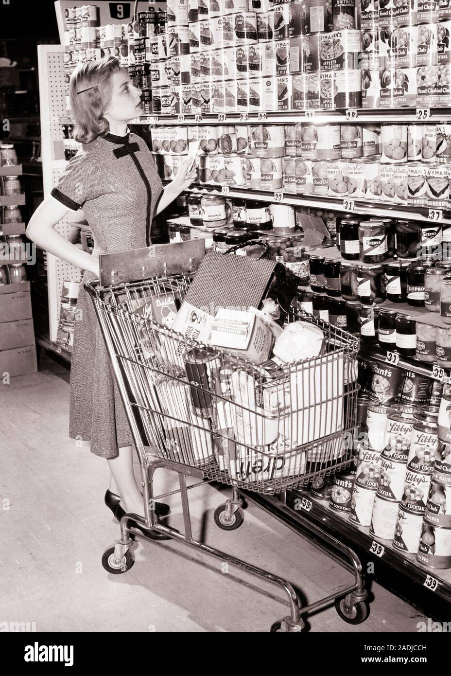 1950s 1960s WOMAN PUSHING SHOPPING CART IN GROCERY STORE HOLDING HER SHOPPING LIST AND LOOKING UP AT SHELVES OF CANNED GOODS - s2146 HAR001 HARS LIFESTYLE FEMALES AISLE HOME LIFE FULL-LENGTH LADIES PERSONS INSPIRATION B&W CANNED SHOPPER GOALS HOMEMAKER SHOPPERS HOMEMAKERS GOODS AND CHOICE AT IN OF UP HOUSEWIVES CONCEPTUAL STYLISH LOOKING UP YOUNG ADULT WOMAN BLACK AND WHITE CAUCASIAN ETHNICITY HAR001 OLD FASHIONED Stock Photo