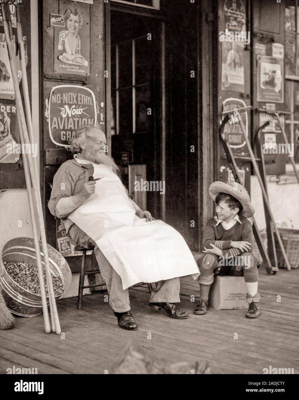 1930s ELDERLY SHOPKEEPER WEARING APRON SMOKING PIPE SITTING WITH YOUNG BOY WEARING STRAW HAT RAGGEDY CLOTHES GENERAL STORE PORCH - s5449 HAR001 HARS STRAW PORCH SENIORS OLD TIME NOSTALGIA OLD FASHION 1 JUVENILE COMMUNICATION TEAMWORK GENERATIONS GRANDFATHER GRANDPARENTS PLEASED FAMILIES JOY LIFESTYLE SATISFACTION ELDER GRANDPARENT PIPE GENERAL RURAL UNITED STATES COPY SPACE FRIENDSHIP FULL-LENGTH HALF-LENGTH PERSONS SHOPS INSPIRATION UNITED STATES OF AMERICA CARING MALES B&W NORTH AMERICA NORTH AMERICAN HAPPINESS OLD AGE OLDSTERS CHEERFUL HIGH ANGLE OLDSTER OWNER TOBACCO GENERAL STORE EXTERIOR Stock Photo