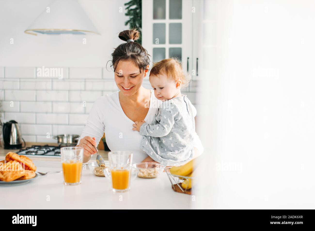 Young woman mom with baby girl on hands cooking breakfast on bright kitchen at the home Stock Photo