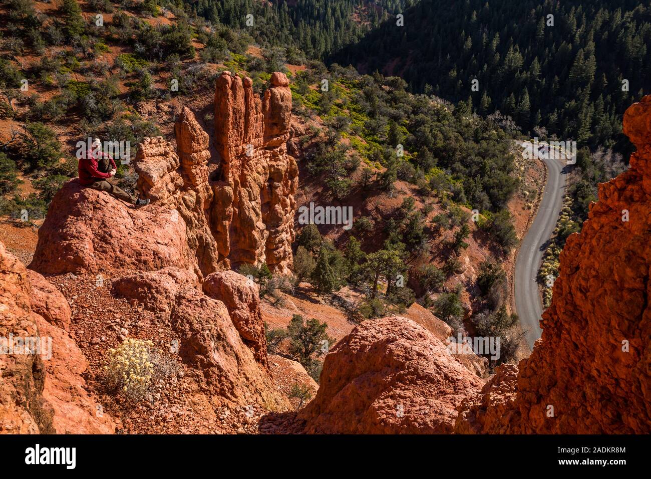 Man sitting atop red rock tower formation above winding canyon road far below. Stock Photo