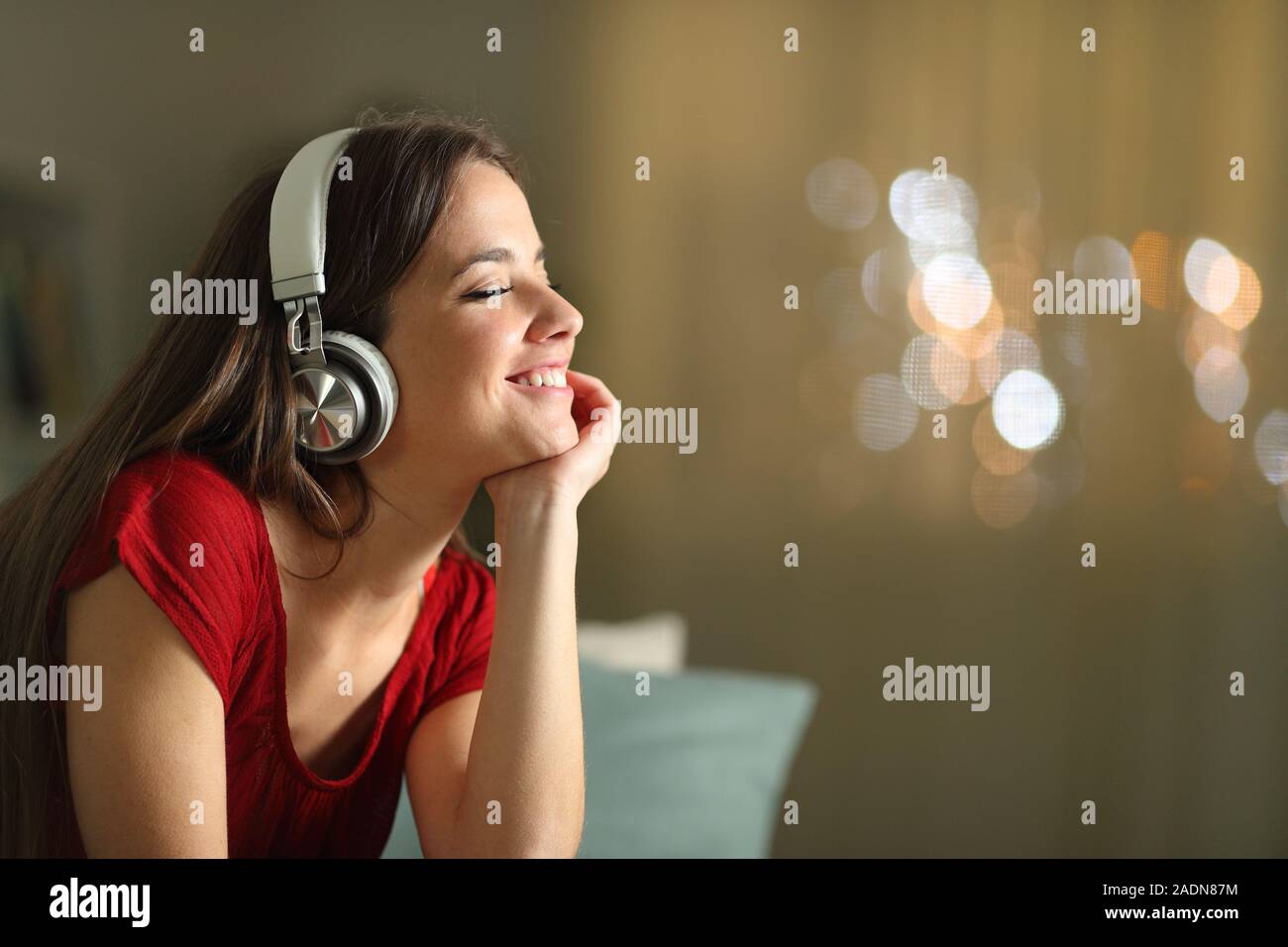 Happy woman wearing wireless headphones listening to music in the night sitting on a couch in the living room at home Stock Photo