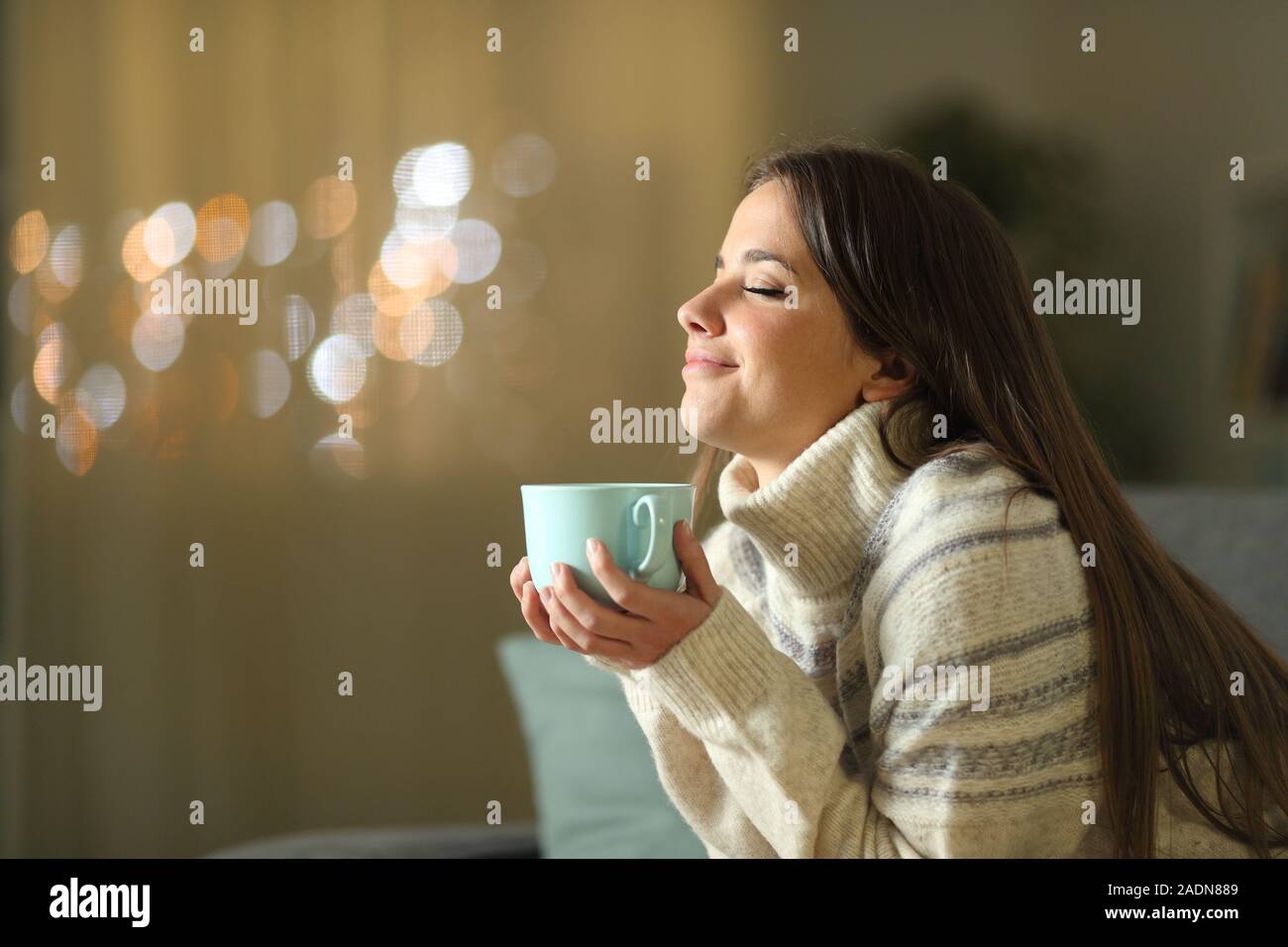 Relaxed woman holding coffee cup in winter sitting on a couch in the living room at home in the night Stock Photo