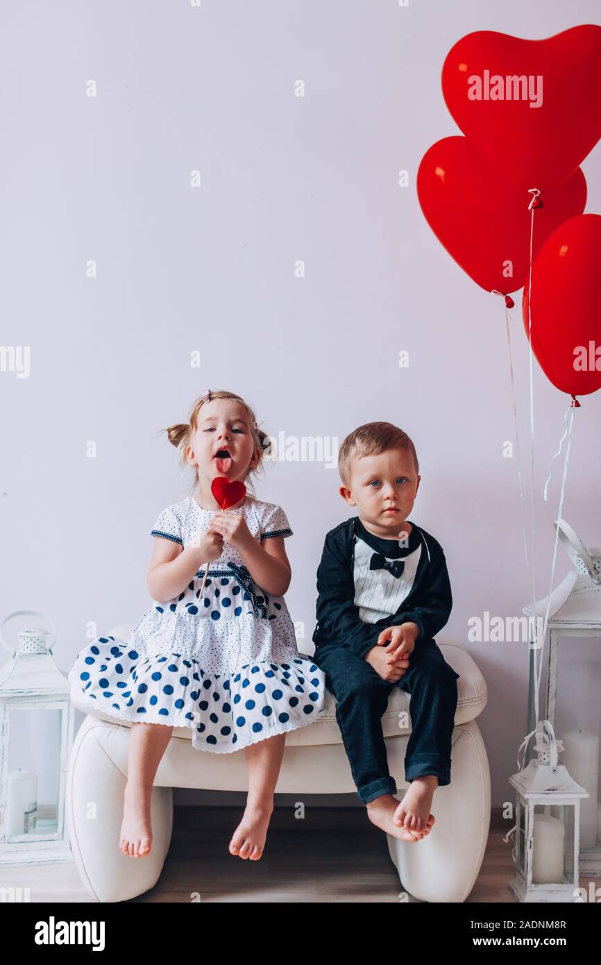 Little girl and boy sitting on a white chair near heart-shaped baloons. Girl licking a red lollipop. Valentines day conept Stock Photo