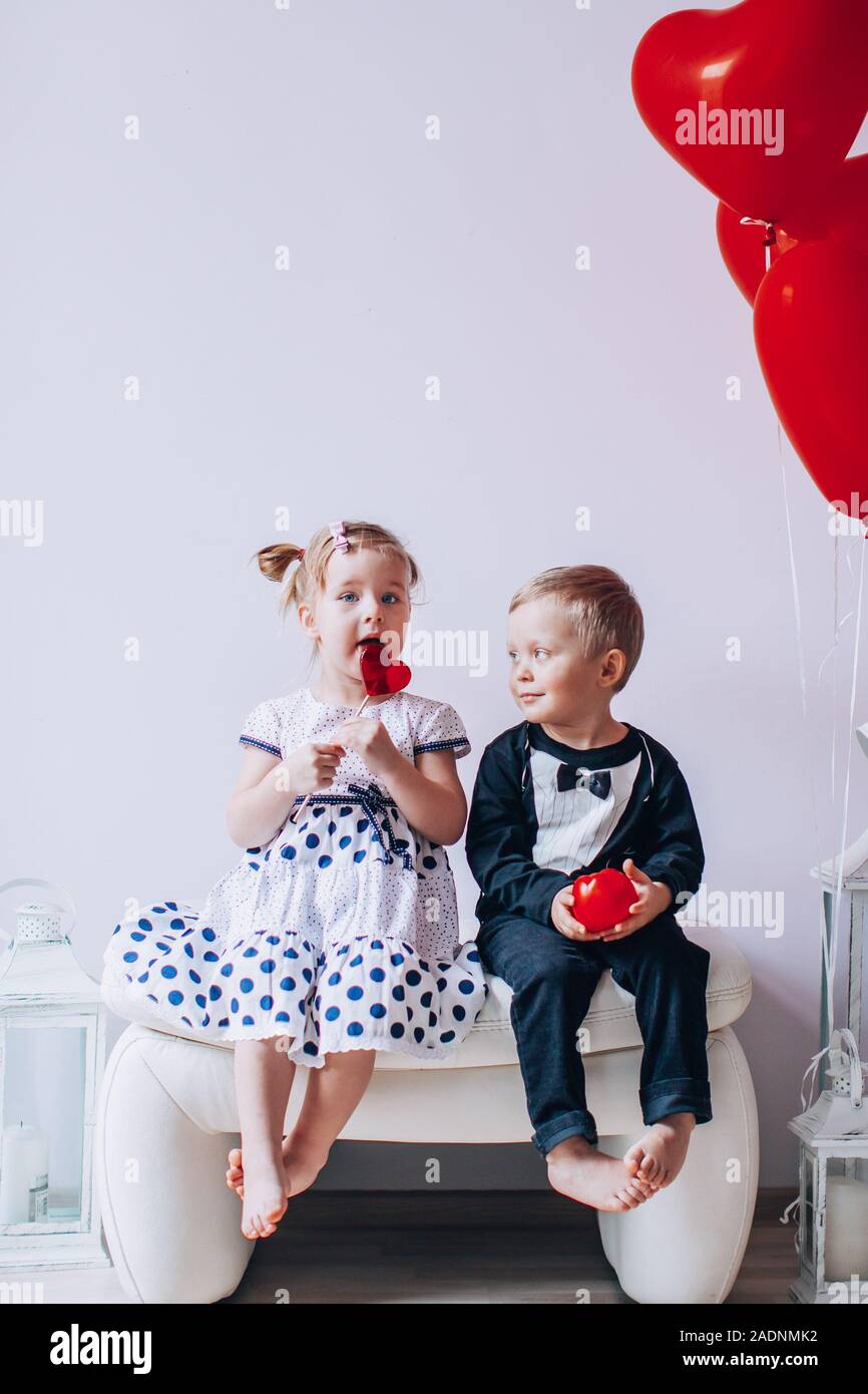 Little girl and boy sitting on a white chair near heart-shaped baloons. Girl licking a red lollipop. Valentines day concept Stock Photo