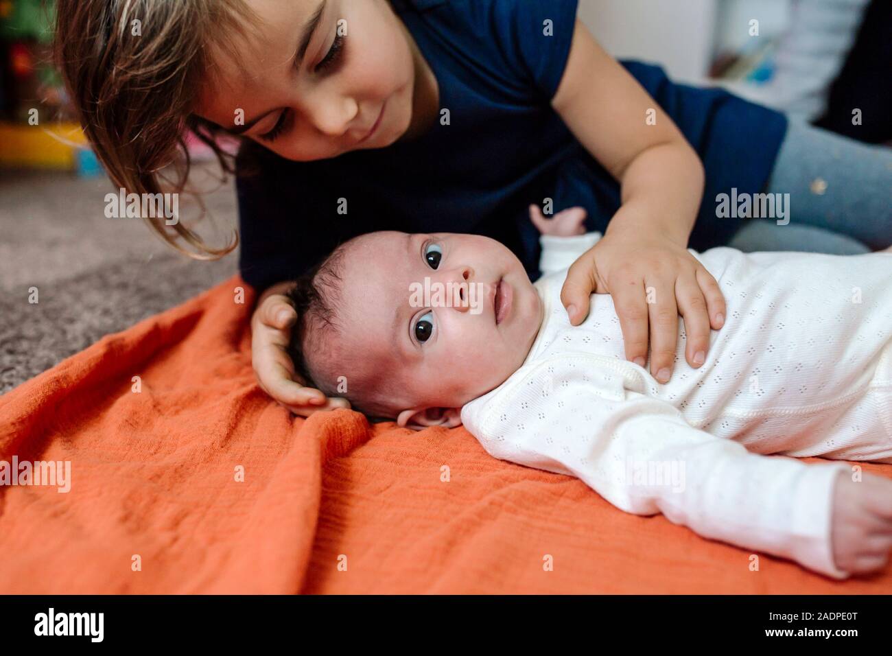 Big sister touching wide-eyed baby with lying on orange blanket Stock Photo