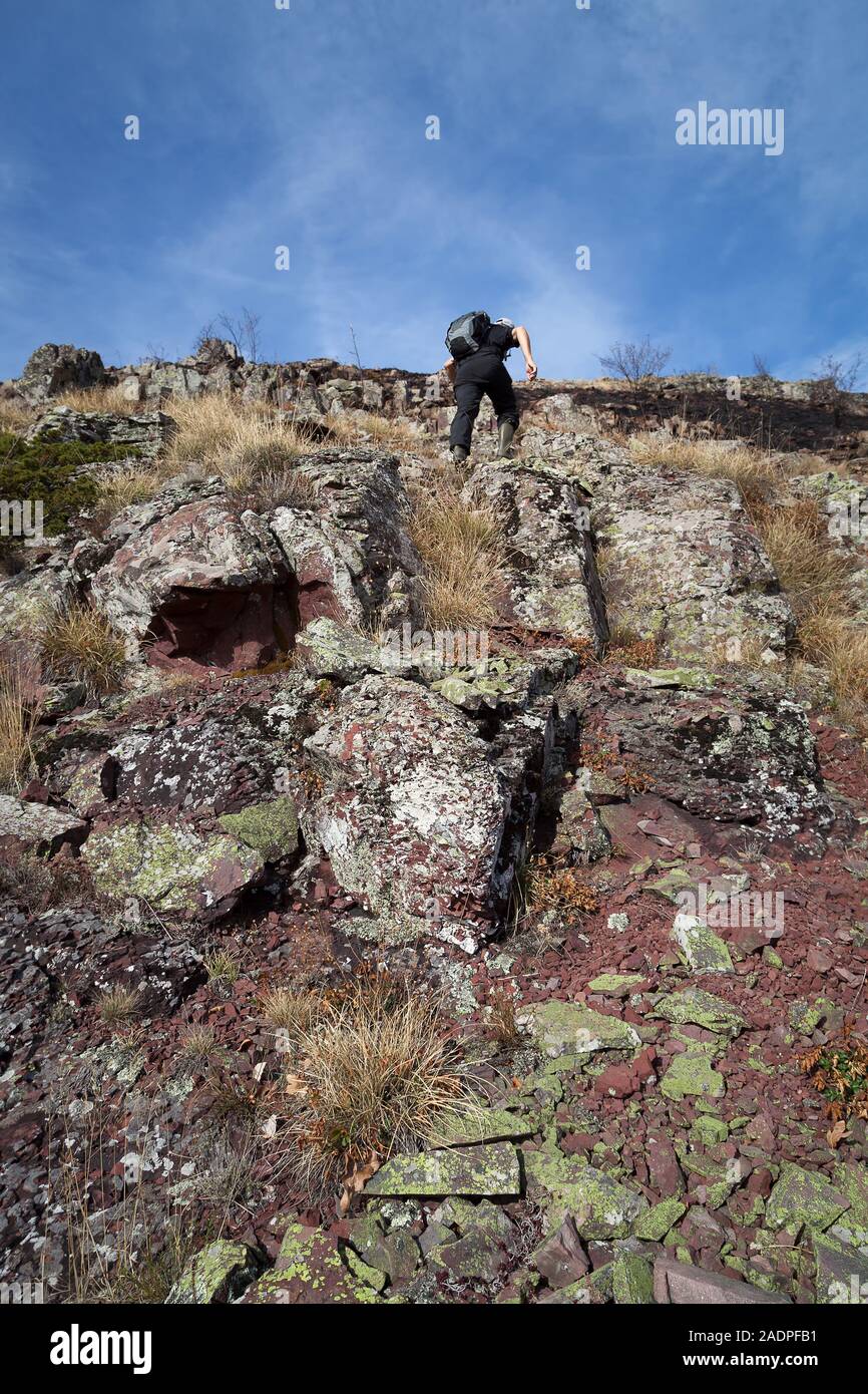 Mountain hiker with backpack and rubber boots, dressed in black, climbing up the steep, rocky, dangerous cliff, burnt by fire and covered by red rocks Stock Photo