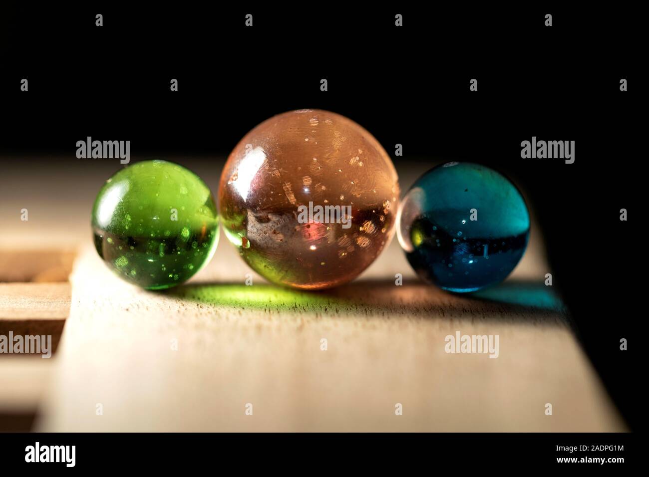 A close up portrait of three different colored marbles on a wooden surface casting colorful reflections. The glass spheres are orange, green and blue. Stock Photo
