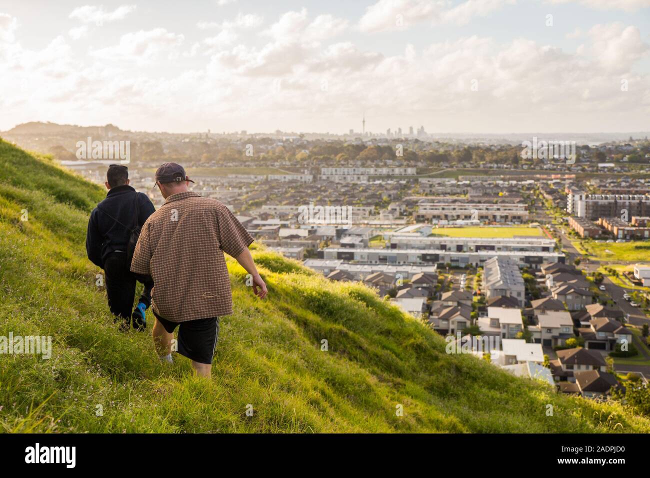 Two males walk on the side of Mount Wellington, overlooking the suburb of Stonefields in Auckland, New Zealand at sunset. Stock Photo