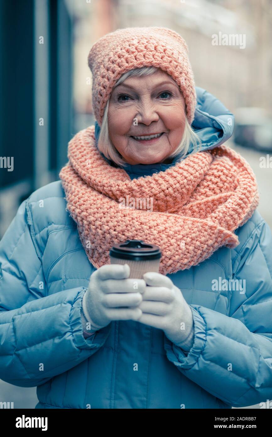 Aged woman looking glad while standing outdoors with coffee Stock Photo